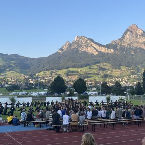 Shinson-Hapkido-Kampfkunst-Show mit Zuschauenden auf Bänken, dahinter auf einem Sportplatz die Vorführenden, die einen großen Kreis machen und in der Mitte eine Pyramide, im Hintergrund von der Abendsonne erleuchtet hohe Berge.