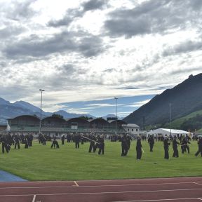 Sportplatz mit Shinson-Hapkido-Schüler*innen in schwarzen Anzügen mit Langstöcken im Vordergrund, dahinter Tribüne und Gebäude vom Sportplatz, im Hintergrund Berge und bewaldete Hügel.
