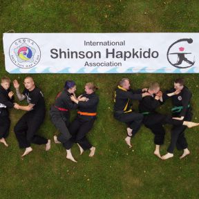Drohnenaufnahme: Eine Gruppe in schwarzen Shinson-Hapkido-Anzügen liegt auf einer Wiese unter einem Shinson-Hapkido-Banner.
