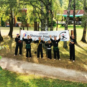 Eine Gruppe in schwarzen Shinson-Hapkido-Anzügen steht winkend unter einem Shinson-Hapkido-Banne.