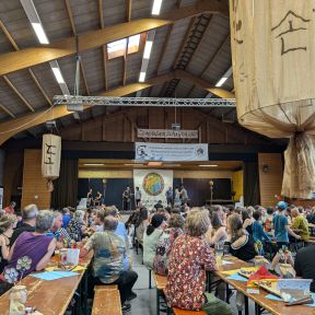 Sporthalle geschmückt mit Shinson-Hapkido-Bannern und großen Lampions, viele Menschen sitzen auf Bierbänken und schauen zu einer Bühne.