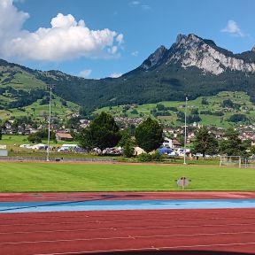 Panorama mit leerem Sportplatz im Vordergrund, leicht ansteigendem Gelände mit Dorf und vereinzelten Häusern im Mittelgrund, im Hintergrund hohe Berge mit bewaldeten Regionen an den Füßen.