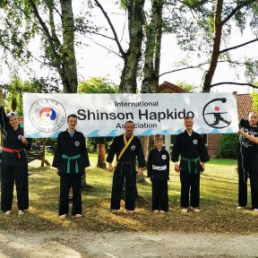 Eine Gruppe in schwarzen Shinson-Hapkido-Anzügen steht lächelnd unter einem Shinson-Hapkido-Banner.