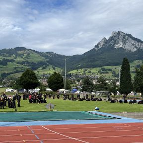 Sportplatz mit Shinson-Hapkido-Schüler*innen in schwarzen Anzügen im Vordergrund, einige mit Langstöcken, vor einem Panorama mit bewaldeten Hügeln und hohen Bergspitzen.
