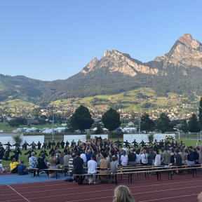 Shinson-Hapkido-Kampfkunst-Show mit Zuschauenden auf Bänken, dahinter auf einem Sportplatz die Vorführenden, die einen großen Kreis machen und in der Mitte eine Pyramide, im Hintergrund von der Abendsonne erleuchtet hohe Berge.