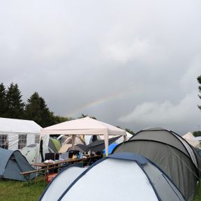 Zeltplatz mit mehreren normalen Zelten und großen Gemeinschaftszelten, am bewölkten Himmel strahlt ein Regenbogen.