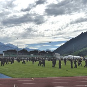 Sportplatz mit Shinson-Hapkido-Schüler*innen in schwarzen Anzügen mit Langstöcken im Vordergrund, dahinter Tribüne und Gebäude vom Sportplatz, im Hintergrund Berge und bewaldete Hügel.